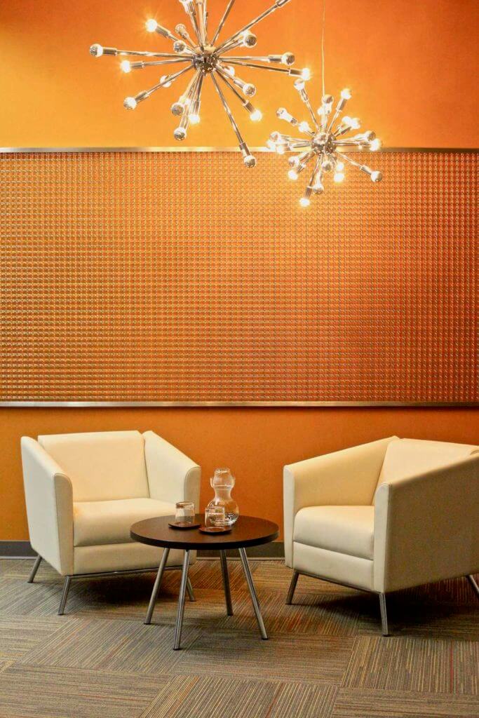 Modern waiting area with silver wall decor, decorative silver chandelier and beige carpet tiles with orange accent color.